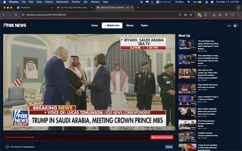 The image depicts a meeting taking place in a formal setting, likely a government or diplomatic venue in Riyadh, Saudi Arabia. In the foreground, there are three men engaged in conversation. 

1. **People**:
   - **Man on the left**: He has light-colored hair and is wearing a dark suit with a white shirt and a blue tie. He appears to be facing the man in the center and is gesturing with his right hand.
   - **Man in the center**: He is dressed in traditional Saudi attire, which includes a white thobe and a black bisht (a cloak) with gold trim. He has a red and white checkered ghutrah (headscarf) secured with an agal (black cord). He is smiling and appears to be engaged in conversation with the man on the left.
   - **Man on the right**: He is wearing a dark suit and has glasses. He is also gesturing with his hands, possibly explaining something to the man in the center.
   - In the background, there are two men in military uniforms, both wearing green berets and adorned with various insignia. They stand at attention, observing the interaction.

2. **Setting**:
   - The background features ornate decor, including a large, framed portrait of a man (not identifiable) on the wall, possibly a Saudi leader. The walls are decorated with intricate patterns, and there are heavy drapes in a dark color hanging from the windows.
   - There are two flags visible: the American flag on the left and the Saudi flag on the right, indicating the diplomatic nature of the meeting.

3. **Text**:
   - At the bottom of the image, there is a banner with the text "BREAKING NEWS" and "TRUMP IN SAUDI ARABIA, MEETING CROWN PRINCE MBS," indicating that this is a news broadcast segment. The logo for Fox News is also visible in the corner.

4. **Overall Context**:
   - The image captures a moment of diplomacy, likely during a significant meeting between U.S. and Saudi officials. The formal attire and the presence of military personnel suggest a serious and respectful atmosphere.

This description includes all observable details without speculation or interpretation.