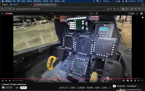 The image depicts the cockpit of a Lockheed F-22 Raptor, showcasing a highly advanced and complex array of controls and displays. 

**Cockpit Details:**
- The cockpit is primarily black, with various screens and control panels arranged in a semi-circular layout.
- There are multiple digital displays, some featuring green and blue graphics, likely indicating flight data, navigation, or targeting information.
- The central display has a radar-like graphic, while others show various symbols and indicators, possibly for altitude, speed, or weapon systems.
- The control panels are bordered with white lights or buttons, which may serve as indicators or controls for the pilot.
- A yellow control handle is visible on the left side of the cockpit, possibly for manual control or emergency functions.
- The seat appears to be designed for a single pilot, with a padded backrest and side supports.

**Background and Environment:**
- The cockpit is situated in a hangar or maintenance area, as indicated by the polished concrete floor and the blurred outlines of aircraft in the background.
- The lighting is bright, suggesting an indoor environment with ample illumination.
- There are no visible people in the image, focusing solely on the cockpit's interior.

**Additional Observations:**
- The cockpit design reflects modern military aviation technology, emphasizing functionality and advanced instrumentation.
- There are no visible logos or branding on the cockpit itself, aside from the inherent design of the aircraft.

This detailed description captures the observable elements of the cockpit without inferring any additional context or intent.