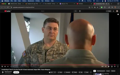 The image shows two individuals engaged in a conversation. 

1. **People**:
   - The person on the left is wearing a military uniform with a camouflage pattern. The uniform has a name tag that reads "BROWN" and an insignia indicating they are part of the U.S. Army. They have short, dark hair and are wearing glasses. Their expression appears serious and focused as they listen to the other person.
   - The person on the right has a bald head and is wearing a light-colored shirt. Their back is facing the camera, making it difficult to see their facial expression. 

2. **Setting**:
   - The background features a wall with a light-colored surface and a window that allows some natural light to enter. The window is partially obscured by a vertical structure, possibly a column or wall feature.
   - There is an American flag visible in the background, indicating a formal or official setting, likely related to military or governmental activities.

3. **Context**:
   - The scene appears to be part of a video segment, possibly an interview or demonstration related to military equipment, as indicated by the title of the video, "Conducting A Test To Try Out The M110 Semi-Automatic Sniper Rifle | Future Weapons."
   - The video is hosted on YouTube, as indicated by the interface at the bottom of the image, which includes options for viewing, subscribing, and interacting with the content.

4. **Text and Logos**:
   - The visible text includes the name "BROWN" on the uniform and the title of the video at the bottom of the screen. The YouTube logo is also present in the top left corner, along with other interface elements typical of the platform.

Overall, the image captures a moment of discussion or instruction in a military context, with a focus on the individuals and their attire, as well as the formal setting indicated by the presence of the American flag.
