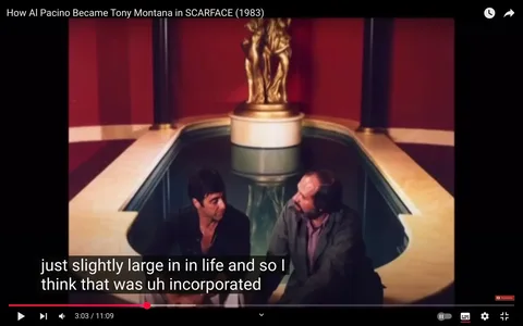 The image depicts a scene from the film "Scarface" (1983). In the foreground, two male characters are seated beside a rectangular pool. The character on the left has dark hair and is wearing a black shirt, while the character on the right has a beard and is dressed in a gray jacket over a plaid shirt. They appear to be engaged in conversation, with the character on the left looking attentively at the other.

The background features a luxurious setting with a deep red wall and a golden statue of two figures, possibly representing a couple, positioned on a pedestal behind them. The pool has a smooth, reflective surface, and the edges are lined with a light-colored stone or marble. The lighting in the scene is warm, contributing to the opulent atmosphere.

The image includes subtitles at the bottom, which provide a transcription of the dialogue being spoken. The text reads: "just slightly large in in life and so I think that was uh incorporated." 

Overall, the scene conveys a sense of intimacy and seriousness, with the characters engaged in a significant discussion within a lavish environment. The visual elements highlight the film's themes of ambition and excess.