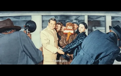 The image depicts a scene with several individuals gathered together, likely at a public event or a photo opportunity. 

**Readable Text:**
- The only visible text in the image is the phrase "Thank you," which is located at the bottom of the image.

**People:**
- In the foreground, a man is prominently featured on the left side. He has short, slicked-back gray hair and is wearing a light-colored coat over a sweater. He is smiling and appears to be shaking hands with a woman next to him.
- The woman next to him is dressed in a dark fur coat and has her hair styled in a bun. She is also smiling and facing the camera.
- Behind them, there are several other individuals. One woman, slightly behind the man, has curly red hair and is wearing a brown fur coat. She is holding a brown clutch bag in front of her face, partially obscuring it.
- Other individuals in the background include a man in a gray hat and coat, and another woman in a lighter coat. Their expressions are not clearly visible due to the angle and distance.
- There are two photographers in the foreground, one on the left with a large camera and the other on the right, slightly obscured by the man in the gray coat.

**Clothing and Appearance:**
- The man in the light coat has a well-groomed appearance and is wearing a sweater underneath. His expression is friendly and welcoming.
- The woman in the dark fur coat has a polished look, with makeup that enhances her features.
- The woman with curly red hair is dressed in a brown fur coat, which appears luxurious.
- The individuals in the background are dressed in various coats and hats, typical of a formal or semi-formal event.

**Setting and Background:**
- The setting appears to be indoors, possibly at an event venue or a terminal, given the white walls and the structure visible in the background.
- The lighting is bright, suggesting a well-lit environment, likely to accommodate photography.
- The presence of photographers indicates that this is a public event, possibly a premiere or a press gathering.

Overall, the image captures a moment of interaction among a group of people, with a focus on the man and woman in the foreground, while the background features additional attendees and photographers.