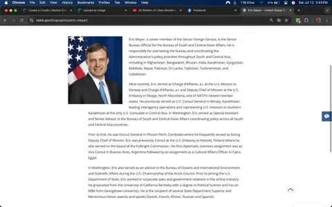 The image features a man in a professional setting, likely an official portrait. He is positioned slightly to the left, facing the camera with a neutral expression that conveys confidence and approachability. His hair is short and neatly styled, and he has a light complexion. He wears a dark suit jacket over a white dress shirt, complemented by a light blue tie, which adds a touch of color to his attire. 

In the background, there is an American flag, prominently displayed, which suggests his affiliation with the U.S. government. The flag's red, white, and blue colors contrast with the darker tones of his suit, drawing attention to both the man and the flag, symbolizing his role in representing U.S. interests.

The setting appears to be a formal office or diplomatic environment, indicated by the professional attire and the flag. The lighting is bright, enhancing the clarity of the image and highlighting the subject's features. 

There are no visible logos or additional text in the image itself, but the context suggests a connection to governmental or diplomatic affairs. The overall tone is serious yet approachable, reflecting the nature of his work in foreign service. The man's expression and posture suggest readiness and professionalism, suitable for someone in a high-level diplomatic role.

This image likely serves to convey trustworthiness and authority, essential qualities for someone involved in international relations. The combination of his attire, the background, and his demeanor all contribute to a sense of professionalism and commitment to service.